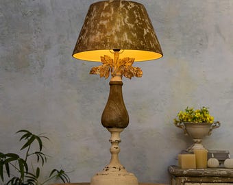 May include: A table lamp with a distressed white and wood base and a gold leaf design. The lampshade is a light brown with a textured pattern.