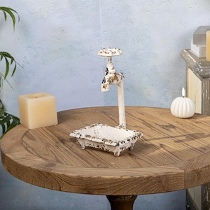 May include: White metal faucet shaped trinket dish with a small rectangular tray. The faucet has a handle and a spout. The dish is sitting on a wooden table.