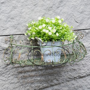 Puede incluir: Un macetero de pared de metal verde con un diseño decorativo de volutas. Una maceta blanca con una pequeña planta verde con flores blancas está sentada en el macetero.