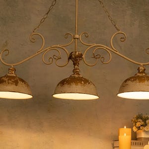 May include: A three-light chandelier with a white metal frame and distressed white and gold finish. The chandelier has a curved design with ornate details and three bell-shaped shades.