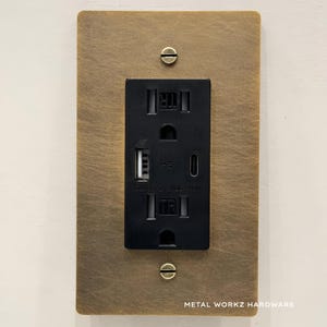 May include: A brushed brass wall plate with a black electrical outlet and two USB ports. The outlet has the text "CLASS 2 125 VAC 15A" printed on it. The plate has the text "METAL WORKZ HARDWARE" printed on the bottom.