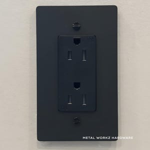 May include: Black metal electrical outlet cover with a standard UK outlet. The cover has a brushed finish and the text "METAL WORKZ HARDWARE" is printed on the bottom.
