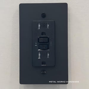 May include: A black electrical outlet cover with a standard three-prong outlet and a GFCI reset button. The cover is labelled "TR" and "METAL WORKZ HARDWARE".