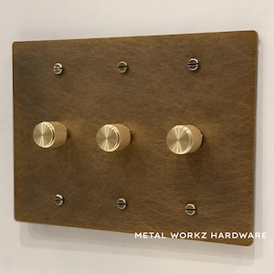 May include: A brass wall plate with three round knobs and six screw holes. The plate has a brushed finish and the text "METAL WORKZ HARDWARE" is printed at the bottom.