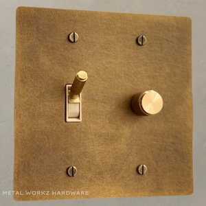 Op de afbeelding: Een messing lichtschakelaar en dimmer met een geborstelde afwerking. De schakelaar en dimmer zijn gemonteerd op een vierkante messing plaat met vier schroeven. De plaat heeft de tekst "METAL WORKKZ HARDWARE" onderaan.