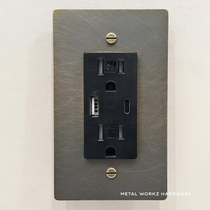 May include: A black electrical outlet with two standard outlets and two USB ports. The outlet is mounted on a brushed metal wall plate with two brass screws. The plate has the text "METAL WORKZ HARDWARE" printed on the bottom.