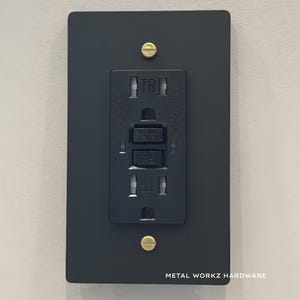 May include: A black electrical outlet cover with a matching outlet. The cover is rectangular with rounded corners and is secured by two brass screws. The outlet has a reset button and is labeled "TR". The text "METAL WORKZ HARDWARE" is at the bottom.