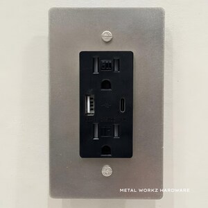 May include: A brushed silver wall plate with a black electrical outlet and two USB ports. The outlet has a standard three-prong configuration. The plate has the text "METAL WORKZ HARDWARE" printed on the bottom.