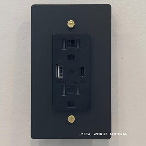 May include: Black electrical outlet cover with a built-in USB charging port and two standard outlets. The cover has a brushed metal finish and gold screws. The text "METAL WORKZ HARDWARE" is printed on the bottom of the cover.
