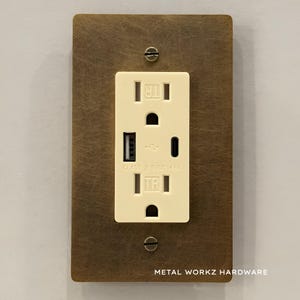 May include: A wall outlet with a USB port and a brown metal faceplate. The outlet is off-white, and the faceplate is rectangular with two screws. The text "METAL WORKZ HARDWARE" is printed at the bottom.