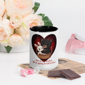 May include: A white ceramic mug with a black interior features a cartoon bear and bunny in a basket inside a heart-shaped design. The mug has the text "Get in the basket. We're going to fall in love."