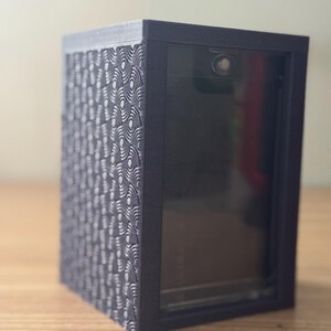 Könnte beinhalten: Eine dunkelgraue Vitrine mit einer transparenten Frontplatte. Die linke Seite der Box weist ein strukturiertes Muster aus sich überlappenden Halbkreisen auf. Die Box steht auf einer Holzoberfläche.