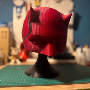 Puede incluir: Una máscara de Daredevil roja y negra con cuernos, expuesta en un soporte negro. La máscara está hecha de un material texturizado y tiene un diseño detallado.