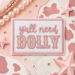May include: A rectangular embroidered patch with a white background and a pink border. The text "y'all need DOLLY" is stitched in pink thread, with "y'all need" in a cursive script and "DOLLY" in a bold, decorative serif font.