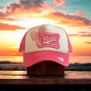 May include: Pink trucker hat with a white mesh back. The front of the hat features a white patch with a pink bow and the text "It's Lake Time".