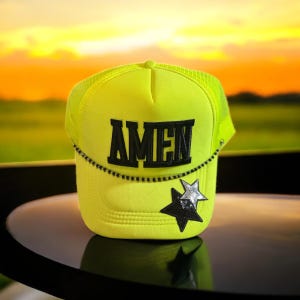 May include: Neon yellow trucker hat with the word "AMEN" in black block letters. The hat features a black beaded chain and two star patches, one black and one silver. The hat is sitting on a black surface.