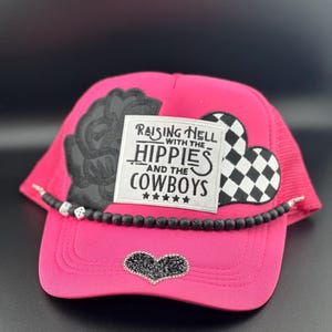 May include: Pink trucker hat with a black rose, a black and white checkered heart, and a black patch that says "Raising Hell with the Hippies and the Cowboys".