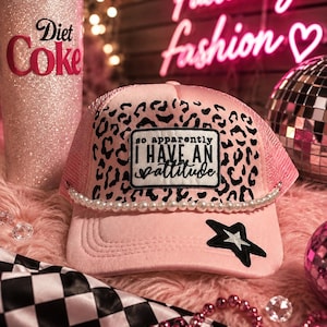 May include: A light pink trucker hat featuring a black leopard print front panel with a rectangular patch that reads, "so apparently I HAVE AN attitude". The hat is accented with a string of white faux pearls and a black star patch on the brim.
