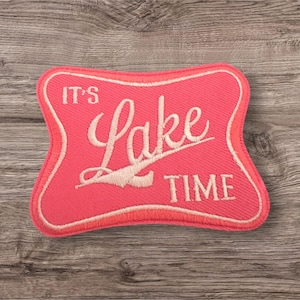 Puede incluir: Un parche bordado de color rosa con costuras blancas que dice "It's Lake Time".