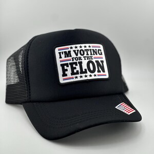 May include: Black trucker hat with a white patch that says "I'M VOTING FOR THE FELON" with red and blue stars. The hat has a black mesh back and a small American flag on the side.