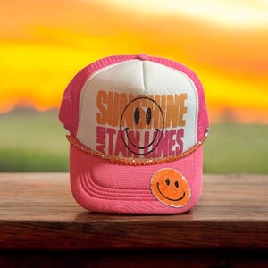 May include: Pink trucker hat with a white front panel featuring the words "SUNSHINE AND TANLINES" and a smiling face graphic. The hat has a pink brim and mesh back, with a small smiley face patch on the front.