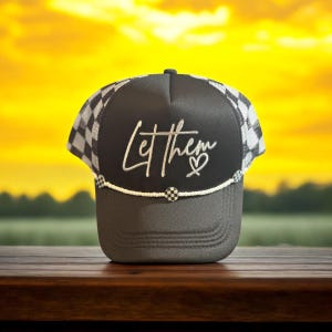May include: Black and white checkered trucker hat with a black mesh back. The hat has a white beaded trim and a silver embroidered design that says "Let Them" with a heart.