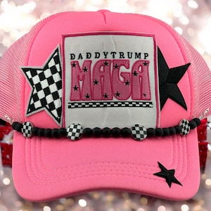 May include: Pink trucker hat with a black and white checkered star, a black star, and a pink patch that says "Daddy Trump MAGA" with stars around the text. The hat has a black beaded trim with white and black checkered beads.
