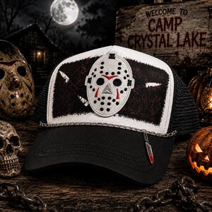 May include: A black and white trucker hat featuring a central embroidered hockey mask patch on a distressed black background. The hat includes a silver-toned metal chain across the brim and a small dangling knife charm with red blood-like accents.