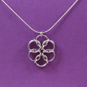 May include: A silver-colored pendant necklace with a floral design. The pendant is made of interlocking circles, suspended from a thin silver chain. The background is a solid purple color.
