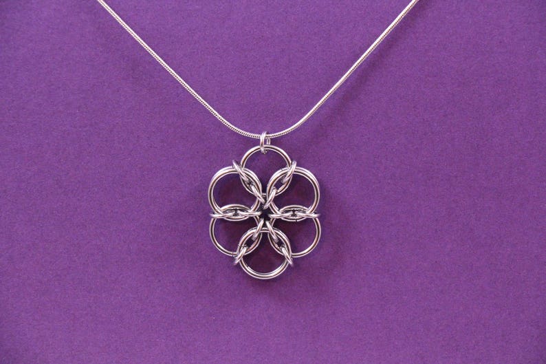 May include: A silver-colored pendant necklace with a floral design. The pendant is made of interlocking circles, suspended from a thin silver chain. The background is a solid purple color.