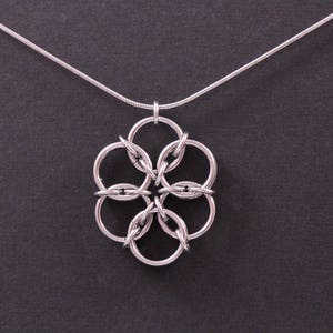 May include: Silver-toned necklace featuring a pendant with a floral design. The pendant is composed of interconnected circular links, creating a flower-like shape. The necklace has a delicate chain and is set against a dark background.