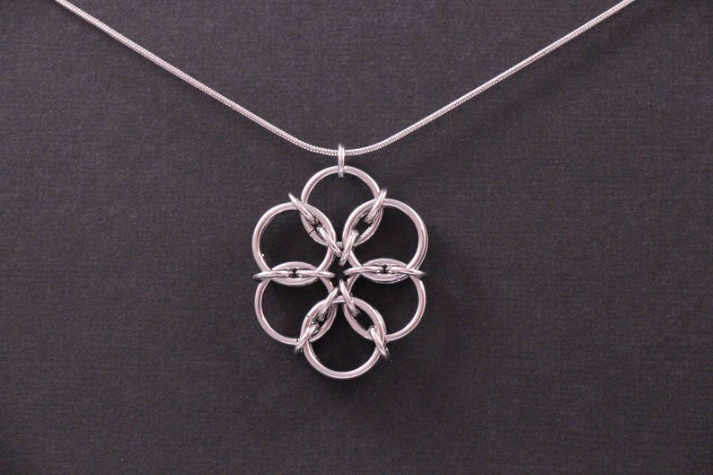 May include: Silver-toned necklace featuring a pendant with a floral design. The pendant is composed of interconnected circular links, creating a flower-like shape. The necklace has a delicate chain and is set against a dark background.