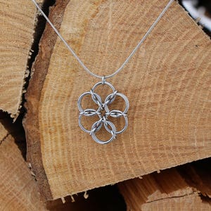 May include: A silver-colored necklace with a flower-shaped pendant. The pendant is made of interconnected circles. The necklace is displayed against a backdrop of stacked, light brown wooden logs, creating a rustic aesthetic.