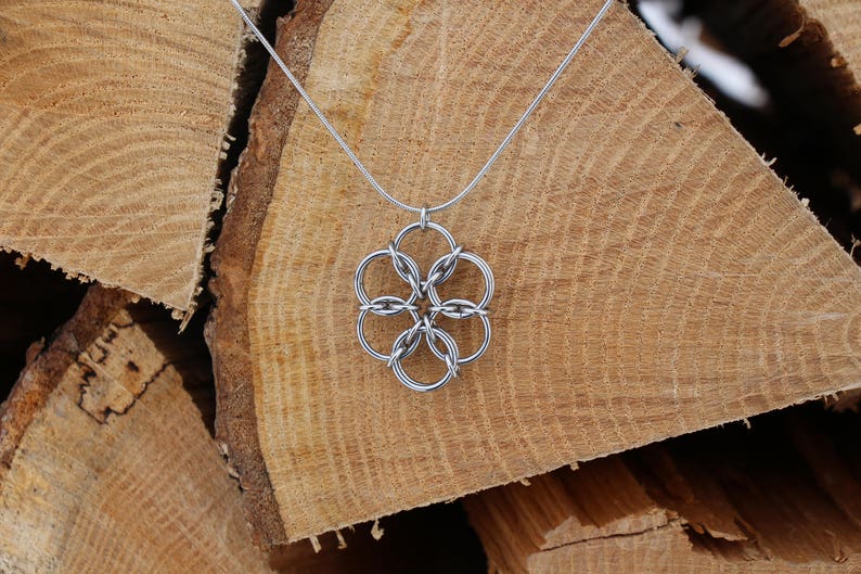 May include: A silver-colored necklace with a flower-shaped pendant. The pendant is made of interconnected circles. The necklace is displayed against a backdrop of stacked, light brown wooden logs, creating a rustic aesthetic.