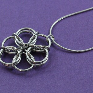 May include: A silver-colored necklace with a flower-shaped pendant. The pendant is made of interlocking rings, creating a detailed floral design. The necklace chain is a thin, snake-style chain. The background is a solid purple color.