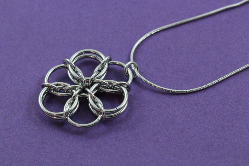 May include: A silver-colored necklace with a flower-shaped pendant. The pendant is made of interlocking rings, creating a detailed floral design. The necklace chain is a thin, snake-style chain. The background is a solid purple color.