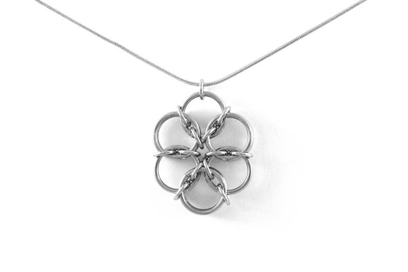 May include: A silver-colored necklace featuring a pendant shaped like a six-petal flower. The pendant is made of interconnected rings, suspended from a delicate chain. The jewelry is displayed against a white background.