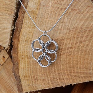 May include: A silver-colored pendant necklace featuring a flower-like design made of interconnected rings. The necklace hangs on a thin, silver chain. The background is a stack of light brown wooden logs, providing a rustic contrast to the jewelry.