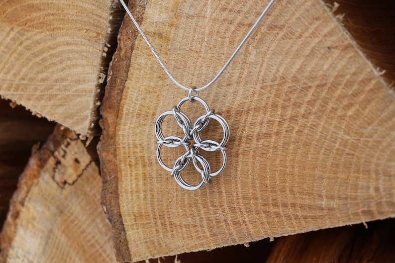 May include: A silver-colored pendant necklace featuring a flower-like design made of interconnected rings. The necklace hangs on a thin, silver chain. The background is a stack of light brown wooden logs, providing a rustic contrast to the jewelry.