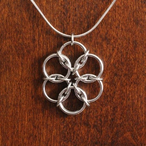 May include: A silver-colored chainmaille pendant necklace. The pendant is a flower-like design made of interconnected rings. The necklace chain is a thin, silver snake chain. The background is a dark brown wooden surface.