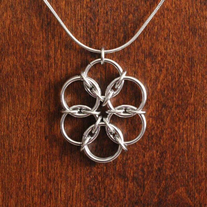 May include: A silver-colored chainmaille pendant necklace. The pendant is a flower-like design made of interconnected rings. The necklace chain is a thin, silver snake chain. The background is a dark brown wooden surface.