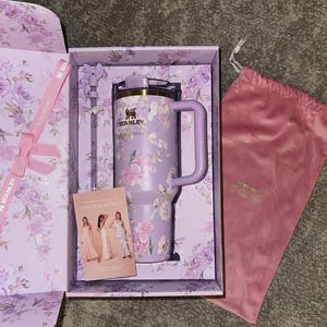 Puede incluir: Un vaso Stanley lavanda con dise&ntilde;o floral, asa y tapa, presentado en una caja a juego con pajita y bolsa rosa con cord&oacute;n. La caja y el vaso presentan el texto "LOVESHACKFANCY".