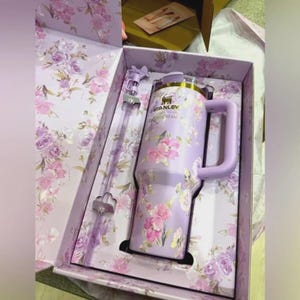 Puede incluir: Un vaso Stanley lavanda con un dise&ntilde;o floral, presentado en una caja de regalo a juego. El vaso tiene un asa y una pajita. La caja y el vaso est&aacute;n adornados con flores rosas y moradas. El logotipo de Stanley es visible.