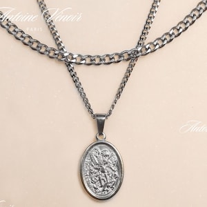 May include: A silver chain necklace with a silver oval pendant featuring a detailed design. The necklace is layered with two chains of different thicknesses.