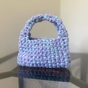 Op de afbeelding: Een kleine, rechthoekige handtas gemaakt van dik, gevlochten stofgaren in een gemêleerd patroon van lichtblauw en zacht lavendel. De tas heeft een enkel, geïntegreerd rond handvat aan de bovenkant en een dichte, gehaakte structuur.