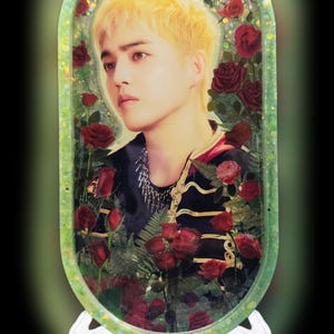 May include: An oval-shaped resin tray with a light green glitter border featuring a portrait of a person with blonde hair surrounded by red roses and green fern leaves. The person wears a black top with gold trim and a silver chain necklace.