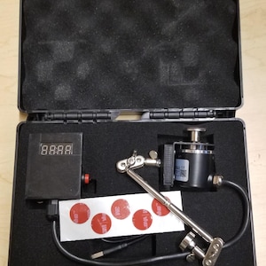 May include: Open black case containing electronic equipment. Includes a digital display showing "88:88", a black cylinder, a silver arm, and several red and white adhesive pads. The case has red latches.