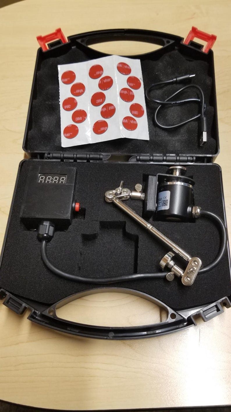 May include: A black carrying case with red catches, open to show tools and components. Includes a digital display, a silver adjustable arm, a black cylinder, and red adhesive circles.
