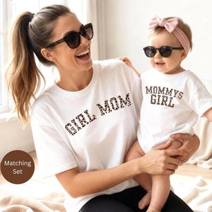 Könnte beinhalten: Weiße T-Shirts mit passendem Leopardenmuster-Text, der "GIRL MOM" und "MOMMY'S GIRL" lautet. Die Shirts werden von einer Frau und einem Baby getragen, beide mit Sonnenbrillen. Das Bild ist ein passendes Set.