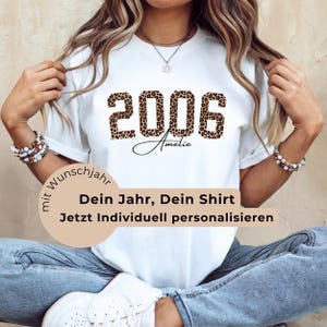 Peut inclure: T-shirt blanc avec l'année "2006" dans un motif léopard. Le nom "Amelie" est écrit en cursive en dessous des chiffres. Le t-shirt contient également du texte en allemand.